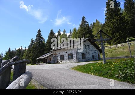Ein weitläufiges Foto im Freien, das ein traditionelles rustikales Steinhaus in einer bergigen Gegend einfängt. Das Gebäude verfügt über Steinmauern und ein dunkles Schindeldach, befindet sich neben einem gepflasterten Weg und einem Holzzaun im Vordergrund, während sich dahinter ein dichter Kiefernwald unter einem strahlend blauen Himmel erhebt. Stockfoto