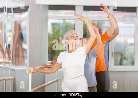 Glückliche, lächelnde alte Frau in Sportbekleidung, die Ballettchoreografie genießt und während des Gruppenkurses im Tanzsalon auf der vierten Position in der Barre stand Stockfoto