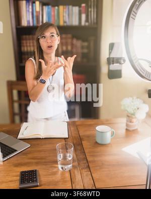 Frau spricht und gestikuliert zu einem Smartphone innerhalb eines Ringlichts an ihrem Heimarbeitsplatz und erstellt Online-Videoinhalte mit einem Laptop, einem Notizblock und einer Tasse. Stockfoto