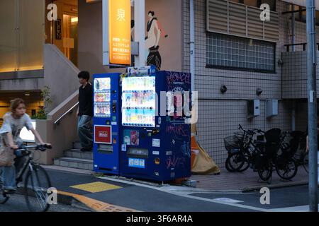 Beleuchtete blaue japanische Verkaufsautomaten vor einem Gebäude mit einer Person, die die Treppe hinuntergeht, und Fahrrädern, die in der Nähe geparkt sind, in Tokio, Japan Stockfoto
