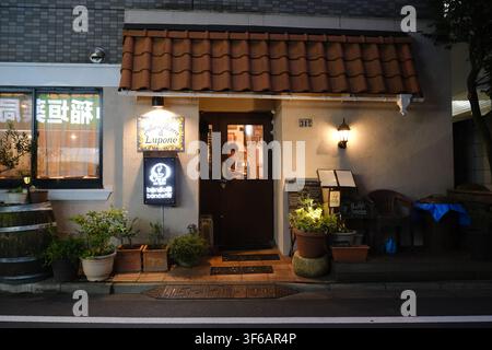 Eine gemütliche italienische Restaurantfassade mit einem Ziegelvordach, beleuchteten Schildern, Topfpflanzen und warmer Beleuchtung in der Dämmerung in Tokio, Japan. Stockfoto