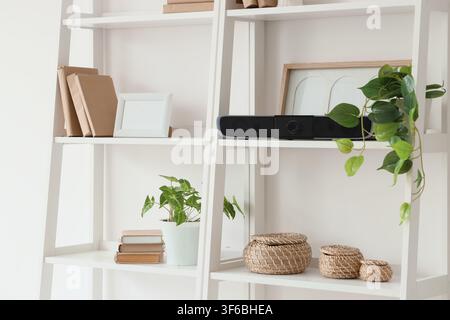 Weißes Holzregal mit Büchern, Topfpflanzen, Soundbar und dekorativen Körben in einem minimalistischen Wohninterieur Stockfoto