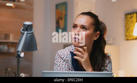 Eine Remote-Mitarbeiterin studiert eine E-Mail auf ihrem Laptop und denkt sorgfältig über die Antwort nach, während sie nach Feierabend an einem Küchentisch sitzt. Stockfoto