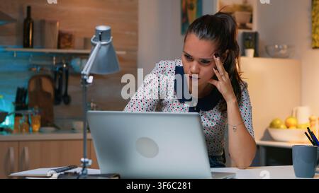 Müde Frau hält ihren Kopf, während sie spät in der Nacht unter dem Druck einer ausgedehnten Aufgabe weiterhin in der Küche am Laptop arbeitet. Stockfoto