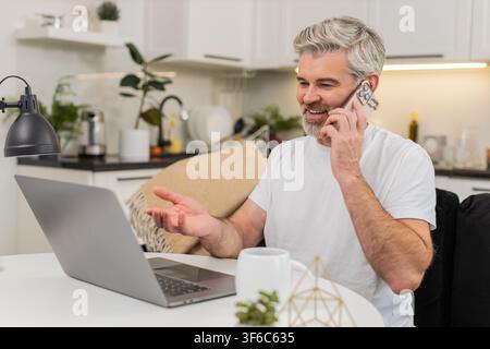 Ein Mann mittleren Alters sitzt zu Hause am Tisch, arbeitet am Laptop und spricht mit dem Kunden über das Smartphone. Reifer Mann auf Sofatypen antwortet, spricht eine ruhige Stimme verwaltet die Aufgabenliste und hält den Fokus unter Last klar und deutlich Stockfoto