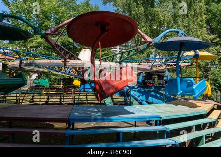 Altes farbenfrohes Karussell mit verwitterten leeren Kabinen steht auf einer hölzernen Plattform, umgeben von grünen Bäumen. Stockfoto