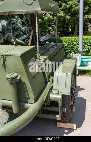 Grüne Militärfeldküche mit Metallabdeckungen und großen Rädern steht auf Asphaltstraße in der Nähe grüner Bäume. Stockfoto