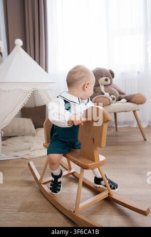 Babyjunge in einem eleganten weißen Hemd und grünen Shorts reitet auf einem hölzernen Schaukelpferd in einem gemütlichen Kinderzimmer Stockfoto