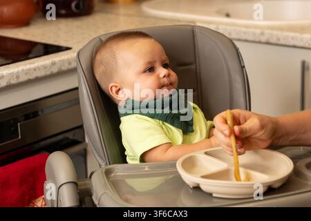 Babyjunge in einem grünen Lätzchen sitzt in einem Küchenhochstuhl mit zusammengekniffenen Lippen, während seine Mutter Essen auf einem geteilten Teller umrührt und einen weiteren Löffel zubereitet. Stockfoto