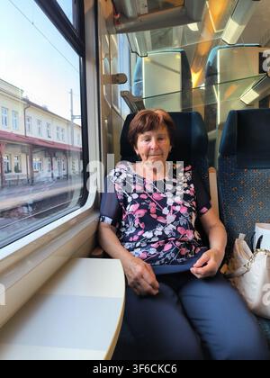 Senior-Frau-Reisende sitzt in einem modernen Zug und schaut aus dem Fenster auf einen Bahnhof Stockfoto