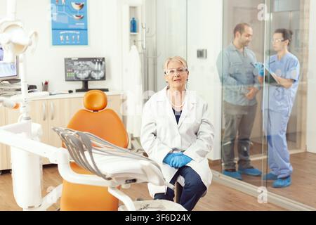 In einer funktionierenden Zahnarztpraxis blickt ein Zahnarzt in die Kamera, während die Behandlung und Empfangspflichten in der Nähe fortgesetzt werden, und das Empfangspersonal die Patientenbesuche in der gesamten Praxis koordiniert. Stockfoto