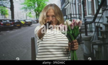 Frau zeigt mit dem Finger, während sie einen Strauß Tulpen auf einer Wohnstraße mit geparkten Autos und Treppenhäusern hält; Einladung zum Selbstvertrauen. Stockfoto