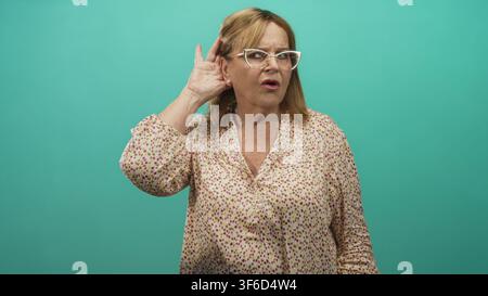 Frau, die Hand an Ohr schröpft und hinter einer Brille in einem Studio-blaugrünen Hintergrund knirscht; Hörschwierigkeiten sind besorgniserregend. Stockfoto