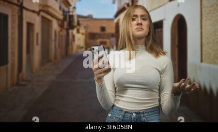 Frau hält ein Smartphone und runzelt die Stirn, während sie mit einer offenen Hand in einer engen Kopfsteinpflasterstraße mit Torbogen und Gebäuden gestikuliert; Frustration digital l Stockfoto