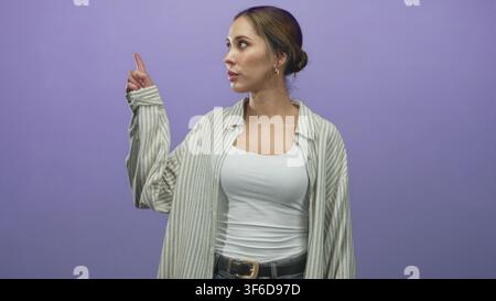 Frau zeigt den Zeigefinger nach oben und Gesten mit der anderen Hand im Studio mit violettem Hintergrund; Neugier-Entdeckung. Stockfoto