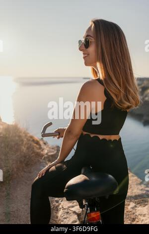 Frau Fahrrad Sonnenuntergang lächelnde Frau sitzt auf ihrem Fahrrad mit Blick auf das ruhige Meer zur goldenen Stunde, während sie eine Sonnenbrille trägt Stockfoto