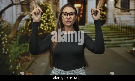 Frau zeigt mit beiden Zeigefingern nach oben auf einen Gebäudeeingang, trägt Brille, lange Haare und Jeans; verspieltes Selbstvertrauen. Stockfoto