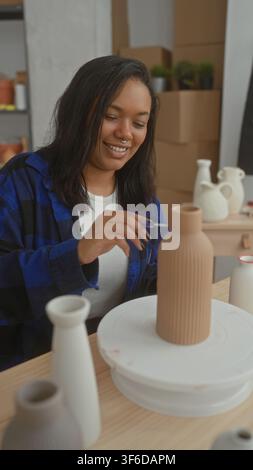 Junge afroamerikanische Frau malt und hält eine Keramikvase auf einer Töpferscheibe im Studio; konzentrierte Kreativität. Stockfoto