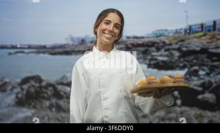 Die Chefin hält ein hölzernes Tablett mit Muffins und zeigt den Finger auf die Kamera auf der Straße an der felsigen Küste mit sichtbarem Pier; freundliche Gastfreundschaft. Stockfoto