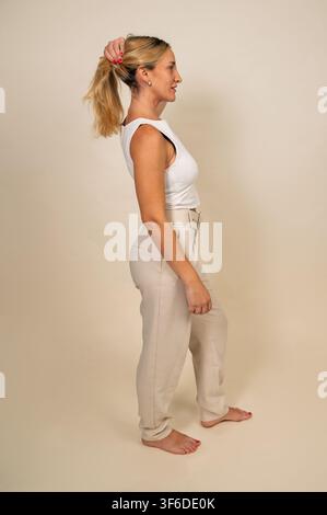 Frau, die ihr blondes Haar zu einem hohen Pferdeschwanz bindet, vor einem schlichten Hintergrund Stockfoto