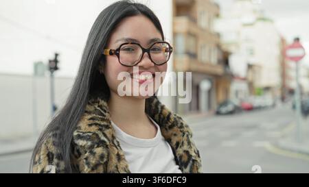 Frau mit Schildpattbrille und Leopardenmantel lächelt mit sichtbarem Gesicht und Ohrring auf einer Stadtstraße; Selbstvertrauen jugendlicher Geist. Stockfoto