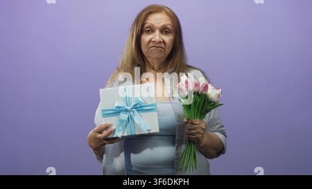 Frau hält eine verpackte Geschenkschachtel mit blauem Band und einen Strauß rosa Tulpen vor einem violetten Studiohintergrund, Blick gesenkt und leichtes Stirnrunzeln; enttäuscht Stockfoto
