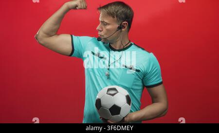 Der Schiedsrichter hält Fußball, trägt Pfeife und Headset, beugt den Bizeps und posiert vor rotem Studio-Hintergrund; Selbstvertrauen. Stockfoto