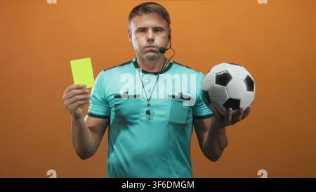 Mann Schiedsrichter hält gelbe Karte und Fußball, Hand hochgehoben mit Pfeife sichtbar im Studio; Autoritätsentscheidung. Stockfoto