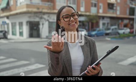 Frau mit Klemmbrett streckt die Hand auf dem Straßenüberweg vor, zeigt und lächelt, Brille sichtbar; freundliche Unterstützung. Stockfoto