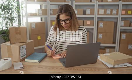 Frau schreibt auf einen Klebezettel neben einem Laptop und gestapelten Kartons im Gebäude; kleines Unternehmen im Fokus. Stockfoto