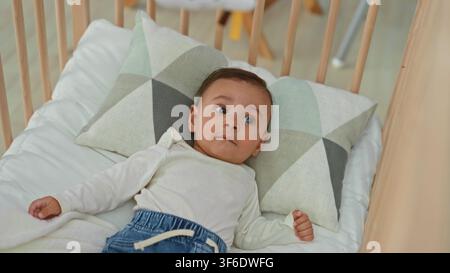 Hispanisches Baby, das in einer Holzwiege auf einem geometrischen Kissen liegt, Hände sichtbar und Kopf gedreht, schaut nach oben in einem Gebäude; ruhige Neugier. Stockfoto