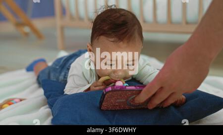 Kleinkind-Junge greift nach einem glitzernden Smartphone, das von einem Erwachsenen gehalten wird, während er auf einer Decke während der Bauchzeit auf einem Beißring kaut; Neugierbindung. Stockfoto