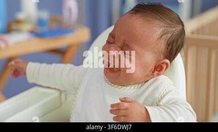 Hispanisches Baby in einem Hochstuhl, das mit dem Löffel gefüttert wird, Essen im Mund und mit verkniffenem Gesicht greifend, weinende Geste in einem Gebäude; Frustrationsverweigerung. Stockfoto