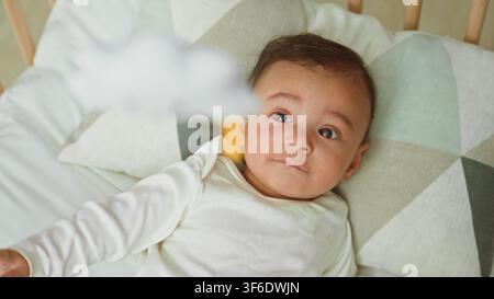 Babyjunge liegt in der Wiege auf geometrischem Kissen und greift nach dem hängenden Mobile über ihm, Gebäude; Neugier verbindet Elternschaft. Stockfoto