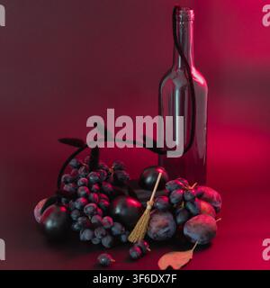 Studio-Halloween-Stillleben mit dunklen Trauben und Pflaumen, arrangiert mit einer Weinglasflasche und einem kleinen Hexenhbesen vor einem tiefroten Hintergrund. T Stockfoto