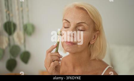 Frau verwendet einen Jade-Gesichtsroller auf Nase und Stirn, während sie auf einem Bett in einem Schlafzimmer sitzt; Selbstpflege-Serenität. Stockfoto