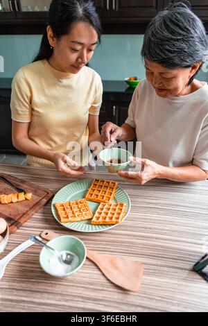 Asiatische Mutter und erwachsene Tochter bereiten zusammen Waffeln in einer heimischen Küche zu und teilen einen warmen Familienmoment, während sie Frühstück mit Sirup und Tischw servieren Stockfoto
