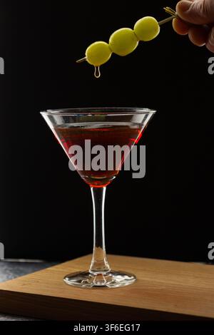 Ein Martiniglas gefüllt mit einem roten Cocktail zeigt rote Trauben, die darin versenkt sind, und grüne Trauben, die auf dem Rand ruhen, vor einem schwarzen Hintergrund auf Stockfoto