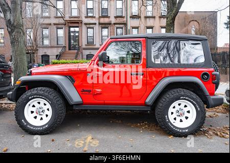 New York, USA - 20. Februar 2026: Roter moderner Jeep Wrangler Offroad-SUV, der am Straßenrand einer Stadtstraße in der Nähe von Wohnhäusern aus braunem Sandstein geparkt ist Stockfoto