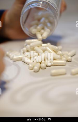 Hand schüttet einen Haufen weißer Medikamentenkapseln aus einer offenen Flasche auf einen Tisch Stockfoto