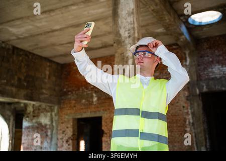 Ingenieur mit Schutzhelm und reflektierender Weste, der sich auf der Baustelle selbst fotografiert. Baustellenmanagement und digitale Kommunikation in Fernberichten Stockfoto