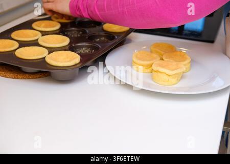 Frau legt frisch gebackene Muffins von einem Tablett auf einen weißen Teller in der Küche Stockfoto