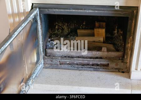 Eine Nahaufnahme des Innenraums eines offenen Kamins, der gestapelte Holzscheite und Aschereste zeigt, wobei die Glastür angelehnt ist Stockfoto