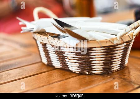 Nahaufnahme eines kleinen geflochtenen Weidenkorbs, gefüllt mit Edelstahlbesteck und weißen Papierservietten, der auf einer rustikalen Holzoberfläche ruht. Stockfoto