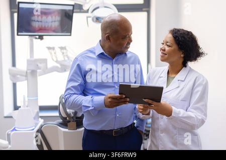 Schwarze Zahnärztin zeigt einem lächelnden älteren männlichen Patienten die Ergebnisse auf einem digitalen Tablet Stockfoto