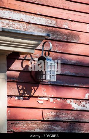 Rostige Laterne hängt an verwittertem rotem Seitenwandverkleidungsbrett. Schatten erstreckt sich über Holz und deutet auf die Wärme des Sonnenlichts hin. Vintage-Charme trifft auf rustikalen Verfall, der an ruft Stockfoto