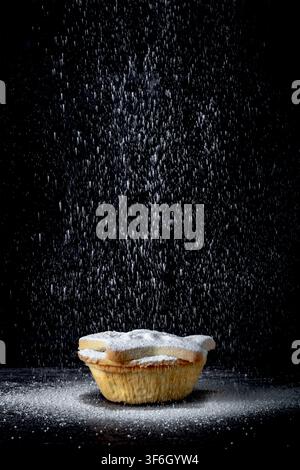 Aktionsaufnahme von Puderzucker, der auf eine einzelne festliche Mince-Pie mit Sternrand fällt, isoliert vor dunklem schwarzem Hintergrund. Weihnachtsessen. Stockfoto