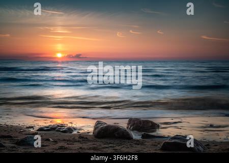 Sanfte Wellen spülen über Felsen an einem sandigen Strand unter einem glühend goldorangefarbenen Sonnenuntergangshimmel in der Dämmerung. Ruhige Meereslandschaft in Morfa Conwy in Nordwales Stockfoto