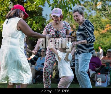 Drei Generationen tanzen gemeinsam bei einem Gemeinschafts-Freiluft-Ereignis in einem öffentlichen Park und feiern Familie, Freundschaft und aktives Leben. Stockfoto