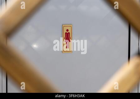 Ein Schild im Retro-Stil, das eine Damen-Toilette anzeigt, ist auf einer hellfarbenen Wand mit einem dezenten Rautenmuster zentriert, umrahmt von verschwommenen Holzelementen Stockfoto